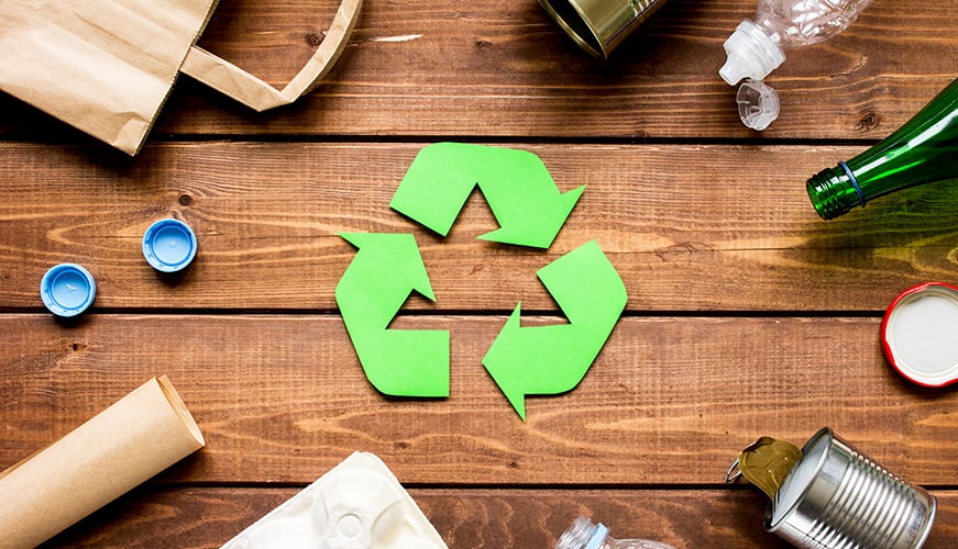 A green recycle symbol on a wooden table surrounded by various recyclable items, including a cardboard tube, bottle caps, glass and plastic bottles, a tin can, and a paper bag.