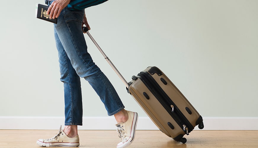 Person walking with a rolling suitcase and holding a passport. Theyre wearing jeans and sneakers, and the suitcase is upright and closed.