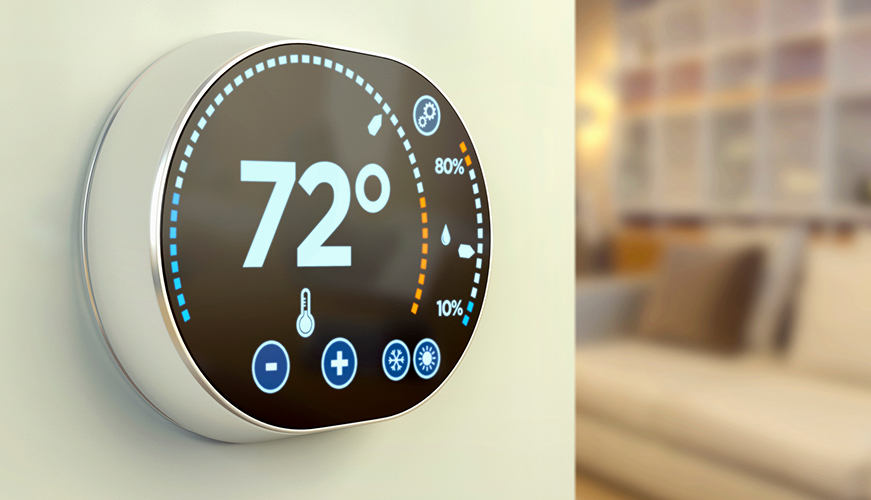 A modern digital thermostat displays a temperature of 72 degrees with blue and orange markers indicating temperature levels. It features plus and minus buttons and a thermometer icon, mounted on a beige wall in a softly lit room.
