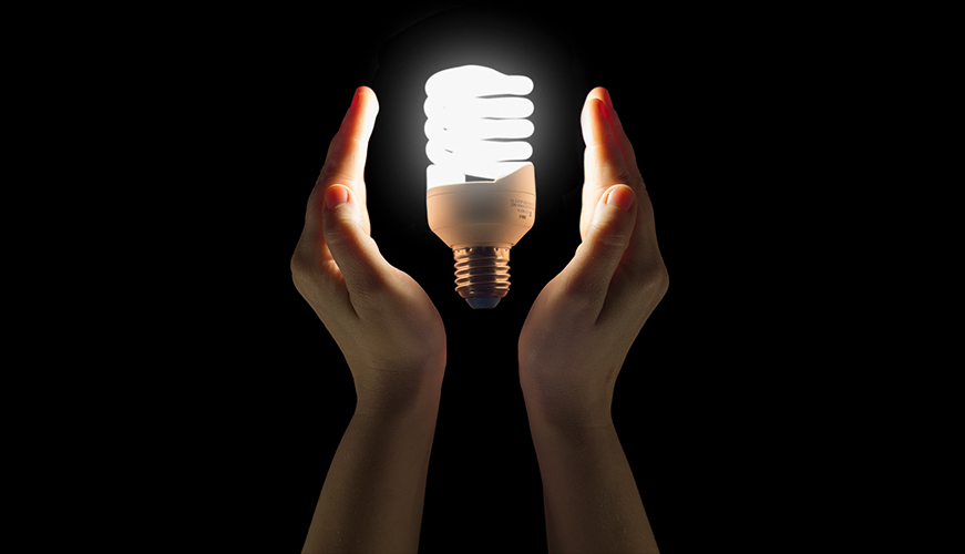 Two hands are cupped around a glowing, spiral-shaped fluorescent light bulb against a dark background.