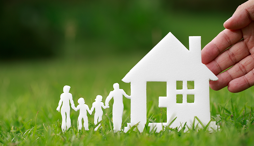 A hand places paper cutouts of a house and a family of four on lush green grass. The scene conveys themes of home, family, and security.