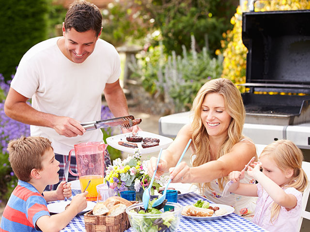 A family of four enjoys an outdoor barbecue. The father grills meat while the mother helps the children at the table. The table is set with food, drinks, and flowers. A barbecue grill is in the background.