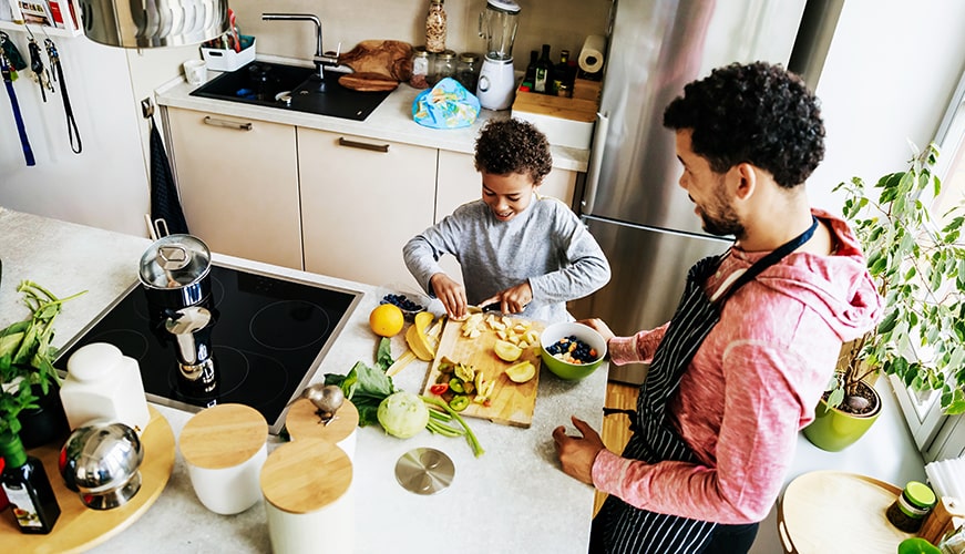 A man and a child are in a kitchen preparing food. The child is cutting vegetables on a wooden board, while the man watches, wearing an apron. The kitchen is bright, with various kitchenware and a plant visible.