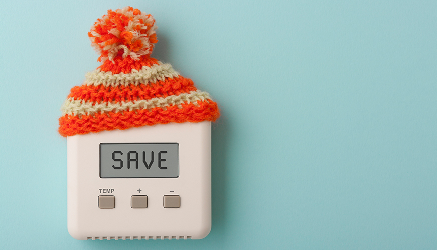 A digital thermostat displaying the word SAVE on its screen is wearing a knitted orange and white woolen hat with a pom-pom, set against a light blue background.