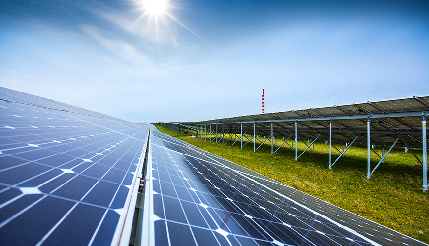 A field of solar panels under a clear blue sky with the sun shining brightly. The panels are mounted on metal frames, and the grass around them is green. A tall red and white striped tower is visible in the background.