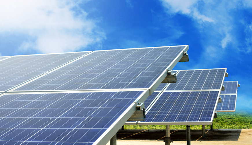 Solar panels arranged in a field under a clear blue sky. The panels are positioned at an angle to capture sunlight, with grass and a partial view of fluffy clouds in the background.