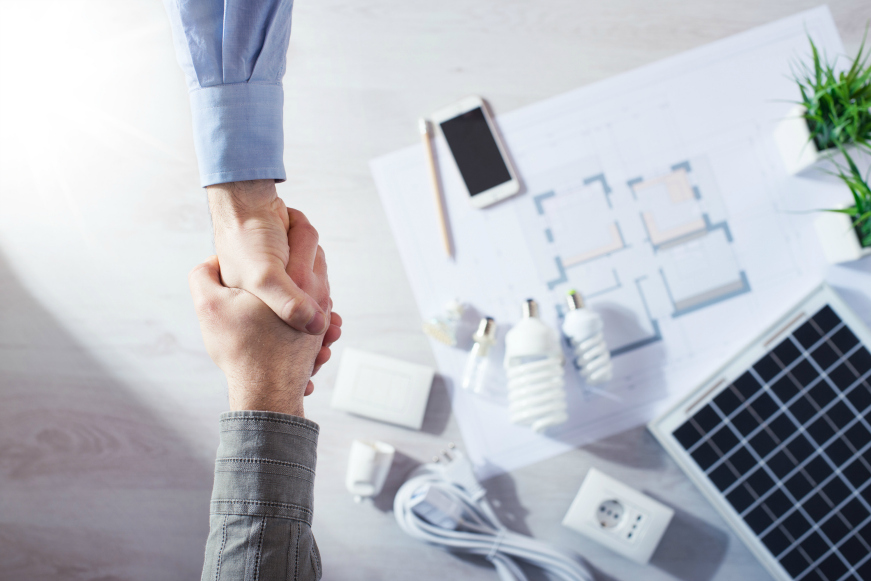 Two people shaking hands over a table with a smartphone, floor plans, energy-efficient light bulbs, a solar panel, and various electrical components on it, suggesting a discussion about sustainable energy solutions.