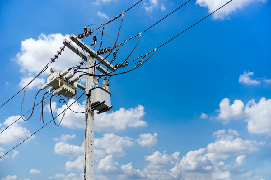 A utility pole with transformers and power lines against a bright blue sky with scattered clouds.