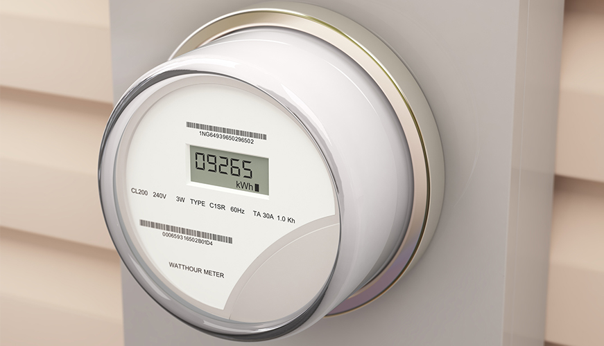 A digital electric meter mounted on a wall displays a reading of 09265 kWh. The device has a circular face with a clear cover, showing text and barcodes. Its installed against a beige siding exterior.