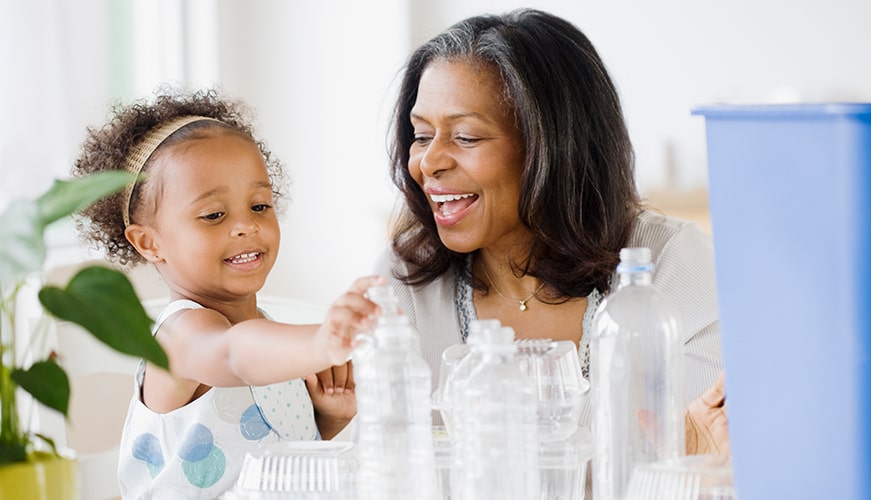 A young girl and an older woman smiling while sorting plastic bottles and containers. They appear to be engaging in a recycling activity indoors, with a plant visible in the foreground.