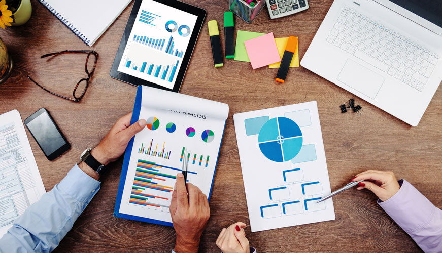 Two people reviewing colorful financial charts and graphs on paper and a tablet, surrounded by office supplies like a laptop, highlighters, and sticky notes on a wooden desk.