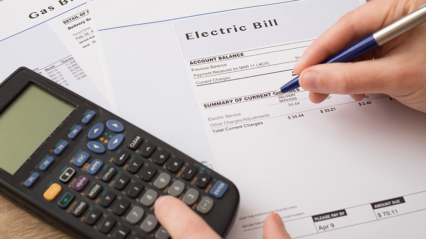 A person holds a pen over an electric bill while using a calculator. The bill details account charges and a balance. In the background, a gas bill is partially visible on the table.