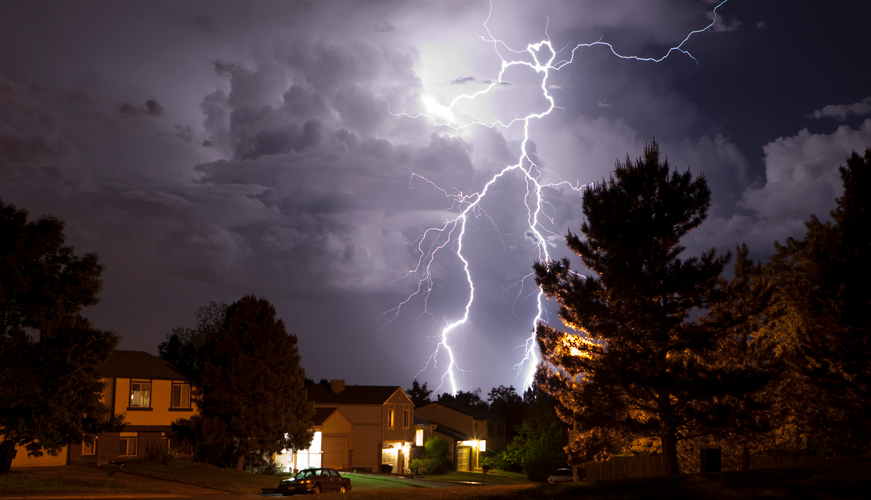 A bright lightning bolt illuminates a cloudy night sky above a suburban neighborhood. Trees and the silhouettes of houses are visible, with several windows lit, creating a dramatic contrast between the stormy sky and the peaceful street.