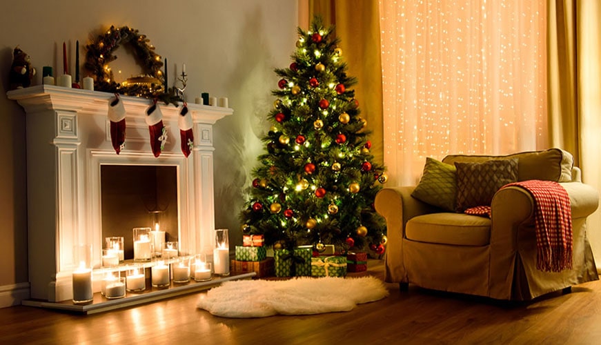 A cozy living room decorated for Christmas with a lit tree adorned with ornaments and lights. A fireplace holds candles and stockings. Gifts are placed beneath the tree, and an armchair with a red blanket sits nearby.