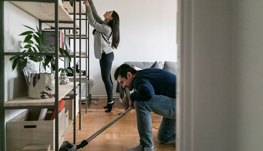 A woman reaches up to adjust items on a tall shelf while a man vacuums the floor in a cozy living room. A potted plant and books are visible on the shelf. The room has a wooden floor and a gray couch.