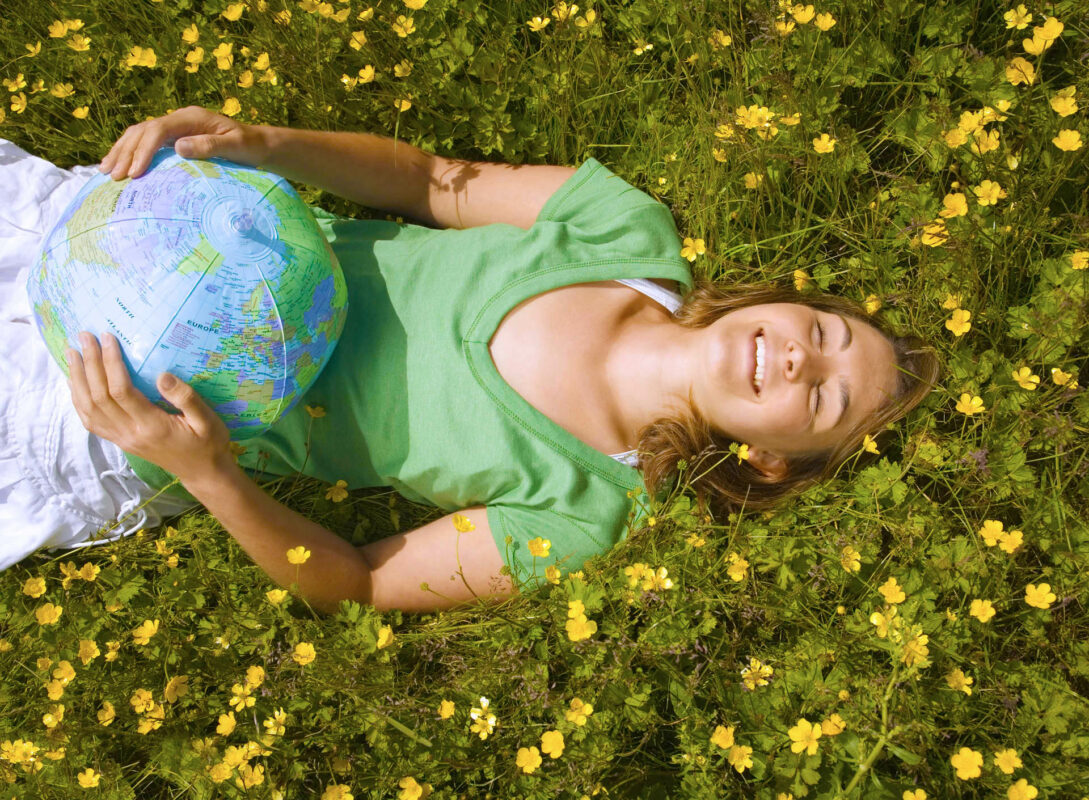A person wearing a green shirt is lying on grass surrounded by yellow flowers, holding a globe and smiling while looking upward.