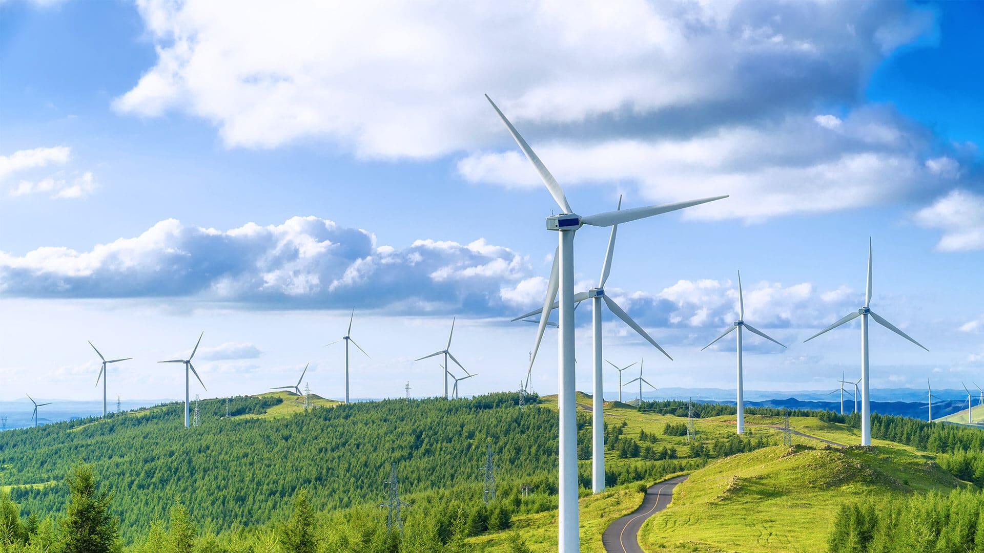 A panoramic view of several wind turbines on a green hill under a partly cloudy blue sky. The wind turbines are surrounded by lush vegetation, and a winding road is visible weaving through the hills.