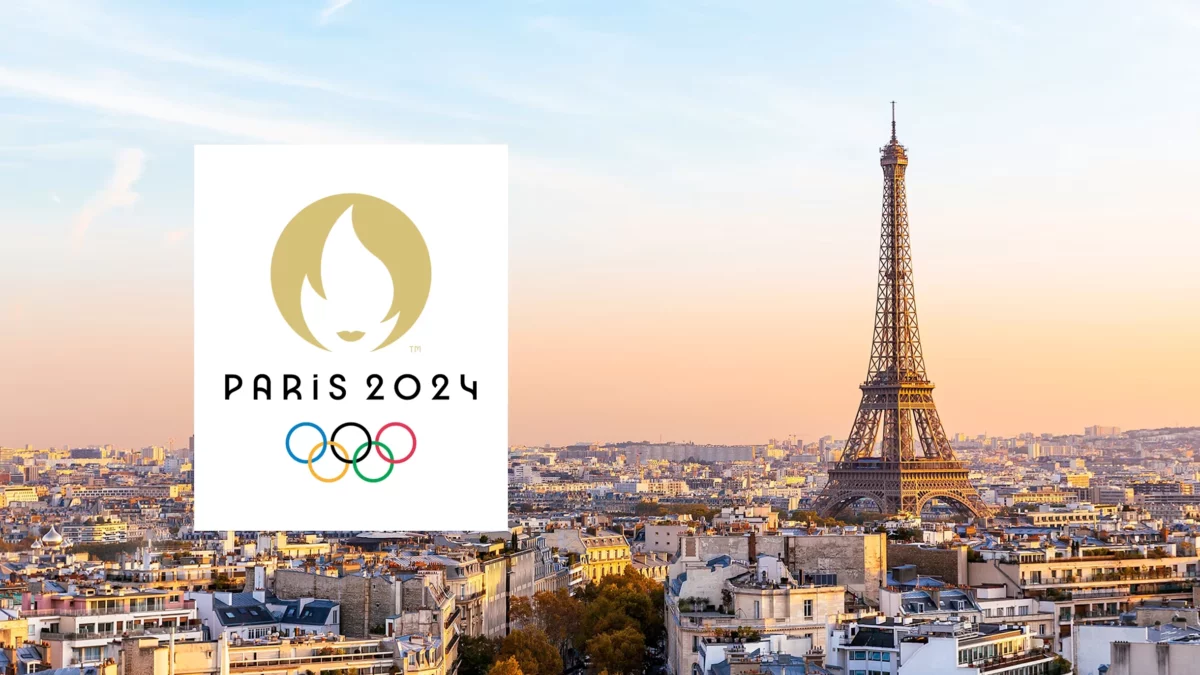 A view of the Eiffel Tower at sunset, with the Paris 2024 Olympics logo on the left featuring a stylized flame and the Olympic rings. The cityscape includes historic buildings under a clear sky.
