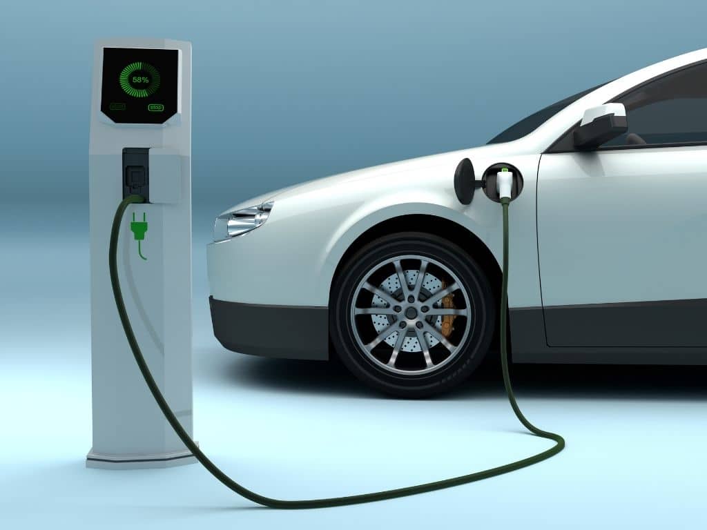 A white electric car is connected to a charging station with a cable. The display on the station shows a charging progress of 65%. The car is parked on a light blue surface with a gradient background.